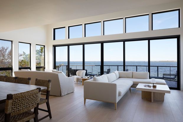 Bright, spacious open-plan living and dining area with expansive black-framed floor-to-ceiling glazing overlooking the ocean. The room features white upholstered sofas and armchairs, a woven dining set, pale timber flooring, and a simple light wood coffee table. A balcony railing and waterfront view are visible beyond the glazing.