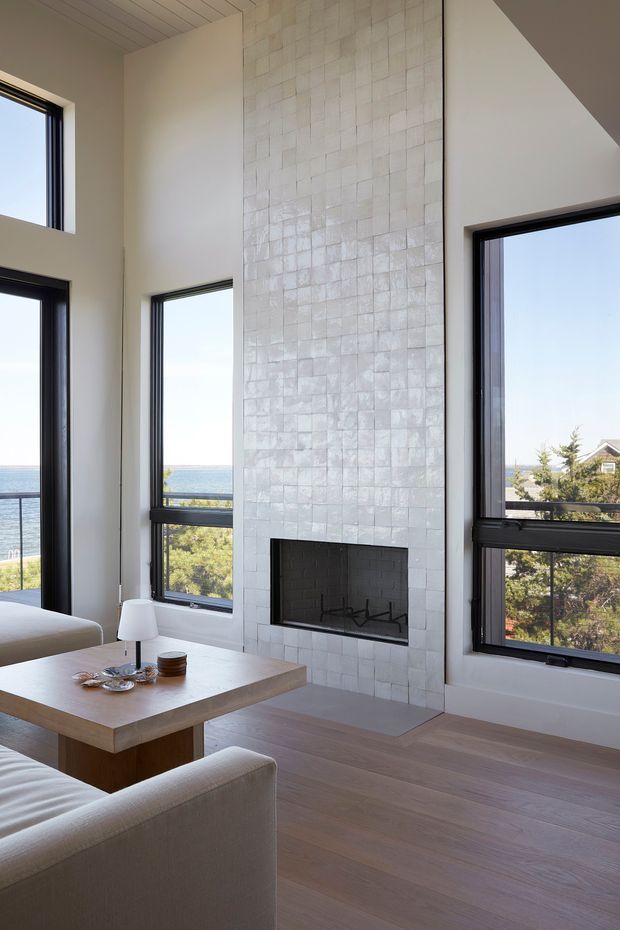 Bright contemporary living room with a tall tiled fireplace feature wall, pale timber flooring, neutral upholstered seating, and a simple wood coffee table. Large black-framed windows provide expansive views toward the ocean and landscaped surroundings. The room has a high, angled ceiling and an airy, minimalist composition.