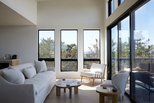 A bright, high-ceilinged contemporary living room with extensive black-framed glazing and views to trees and water beyond. The room features a large light-grey sofa, sculptural upholstered lounge chairs, simple round timber coffee tables, pale walls, and timber flooring. Strong natural light and the vertical windows create an airy, refined atmosphere.