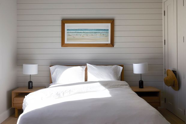 A bright, compact bedroom with a neatly made white bed centered against a horizontal grey-painted shiplap feature wall. Matching bedside tables and white cylindrical lamps flank the bed, while a framed beach artwork adds a coastal accent. Built-in white wardrobe cabinetry is visible on the right, with a woven hat hanging beside it.