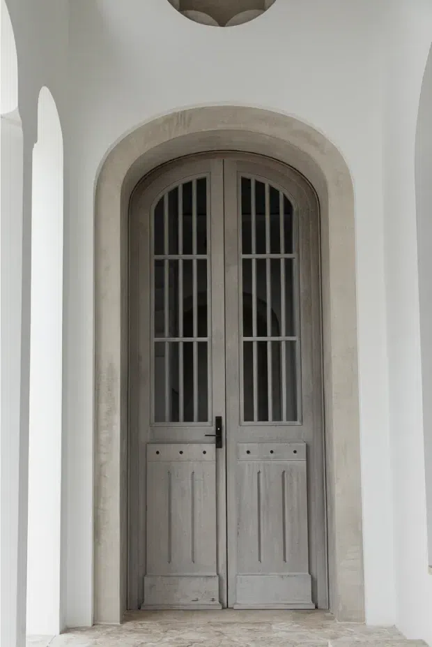 A tall arched double-leaf timber door with narrow vertical glass panels and decorative muntins, set within a deep, textured stone or plaster architrave. The door is framed by white plaster walls with slender arched openings, creating a formal, historic-inspired architectural entrance.