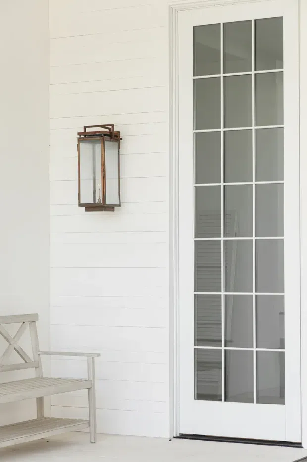 A bright, restrained exterior entry detail featuring a white horizontal weatherboard wall, a tall white multi-pane glazed door, and a traditional bronze wall lantern. A pale timber outdoor bench is partially visible at the lower left. The composition is clean and softly lit, with the door and facade dominating the frame.