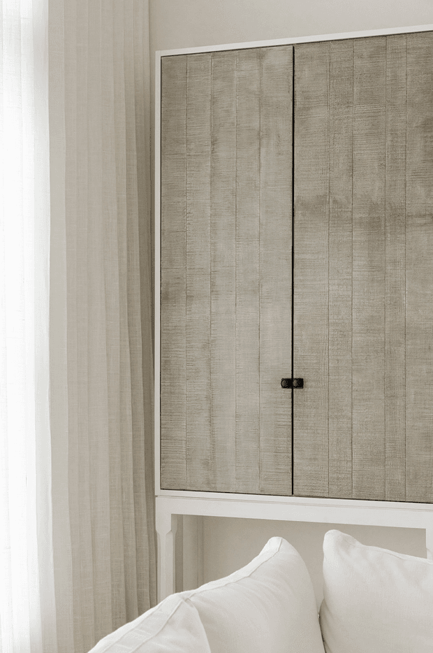 Close-up of a softly styled bedroom corner featuring a large built-in wall cabinet or headboard storage unit with textured taupe-beige doors, slim dark hardware, and white trim. White pillows and bedding sit below the joinery, while sheer neutral curtains filter daylight at the left. The overall appearance is calm, refined, and monochromatic.