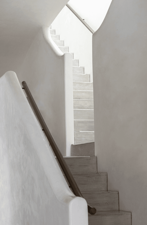 A minimalist interior stairway with pale textured plaster walls, softly rounded parapets, light grey stone or concrete steps, and a dark timber handrail. The stair rises through a narrow, sculptural opening toward a bright upper level, creating a calm and softly lit architectural composition.