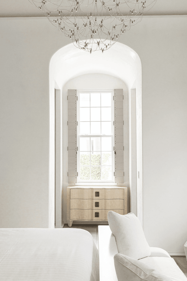 Bright, refined bedroom interior viewed toward a recessed arched window alcove. The room features white walls and bedding, a pale upholstered chair, a light wood chest of drawers beneath the window, narrow built-in shutters or cabinetry flanking the window, and an ornate oversized wire chandelier.