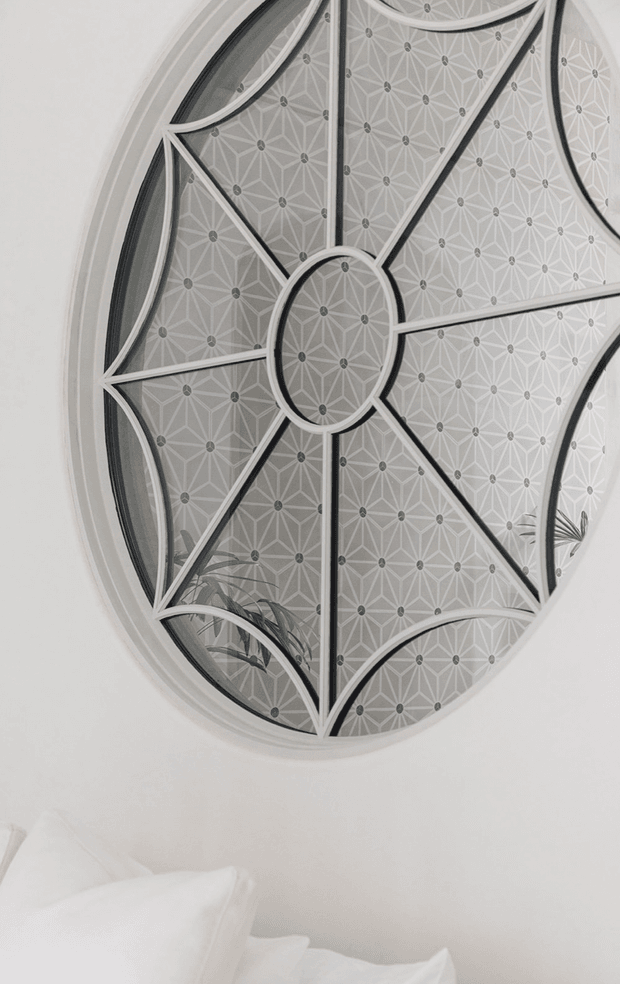 Close-up interior view dominated by a large oval decorative wall mirror with a geometric, radial metal framework and patterned translucent or reflective panels. The mirror is mounted on a white wall above the corner of a white upholstered bed with white pillows. The restrained monochromatic palette and ornate geometric detailing create a refined, lightly art-deco-inspired appearance.