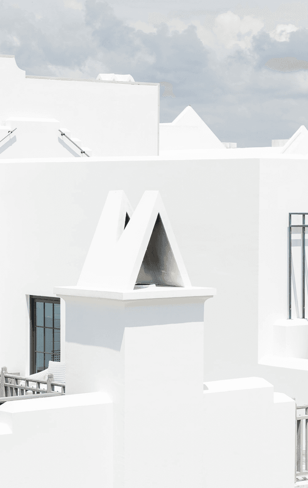 A high-key architectural exterior photograph dominated by white geometric masonry forms, parapets, stepped rooflines, and a sculptural triangular chimney or roof vent. Dark metal-framed windows and railings provide restrained contrast, while a cloudy sky forms the background. The composition emphasizes minimalist, sculptural massing and strong sunlight.