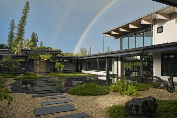 Exterior view of a contemporary multi-level residence arranged around a landscaped courtyard garden and reflective pond. The building features extensive dark-framed glazing, pale rendered walls, a projecting upper level with exposed structural beams and deep eaves, and a low connecting wing. Stepping stones cross gravel and mossy planting beds toward the house, with a rainbow visible in the sky.