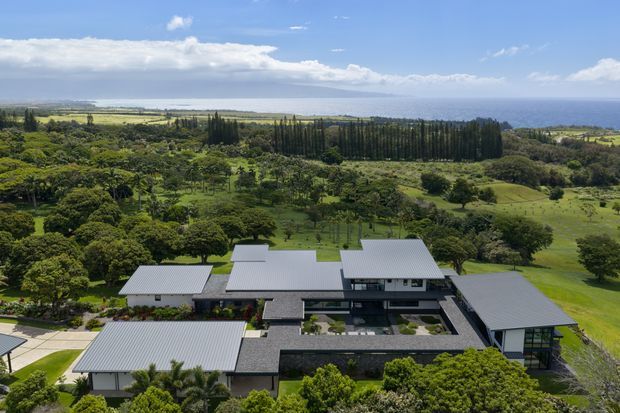 Aerial view of a large contemporary single-storey residential property with multiple low-pitched grey metal roofs arranged around landscaped courtyards. The home is set within expansive green grounds, with mature trees, agricultural land and a visible ocean horizon beyond.