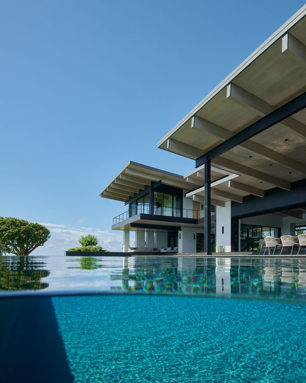 Contemporary multi-level residence viewed from the edge of a turquoise infinity pool. The home features broad cantilevered flat roof planes, exposed dark structural beams, white rendered columns, extensive glazing, balconies with glass balustrades, and an open outdoor dining area. The pool dominates the foreground, reflecting the house and clear blue sky.