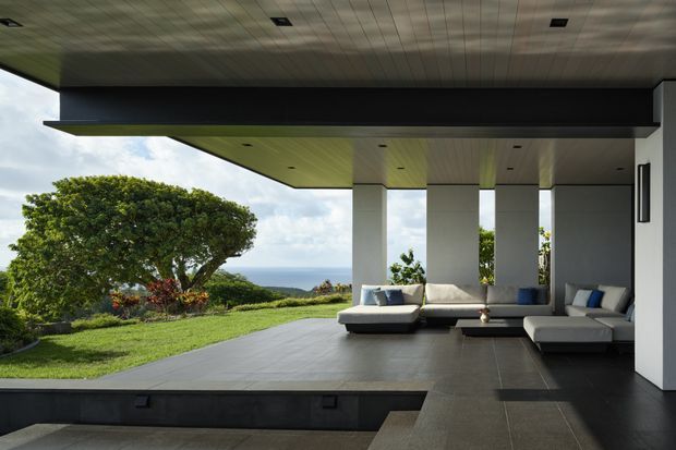 A covered outdoor lounge beneath a dramatic flat-roofed architectural overhang, with white structural columns, dark tiled flooring, and built-in-style light seating arranged around a low table. The open-sided space overlooks a manicured lawn, trees, and a distant ocean horizon. The design is clean-lined and contemporary, with strong horizontal geometry and a restrained black, white, and grey palette.