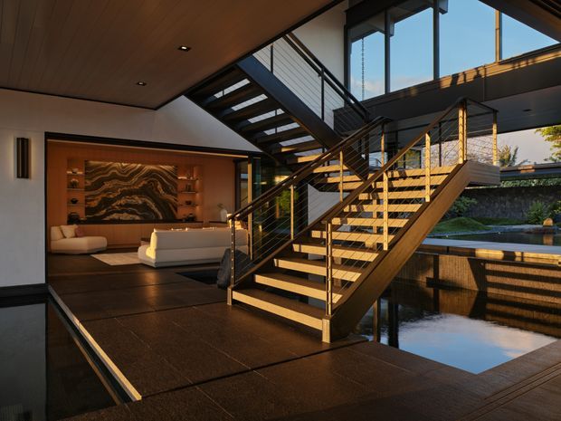 A dramatic contemporary interior centered on an open-riser timber staircase with dark metal framing and cable balustrades. The stair connects a lower living area with a mezzanine or upper level, surrounded by extensive floor-to-ceiling glazing. Dark stone or tile flooring, warm timber ceiling panels, and a textured feature wall frame a sunken lounge, while a reflective water feature or pool is visible beside the circulation space.