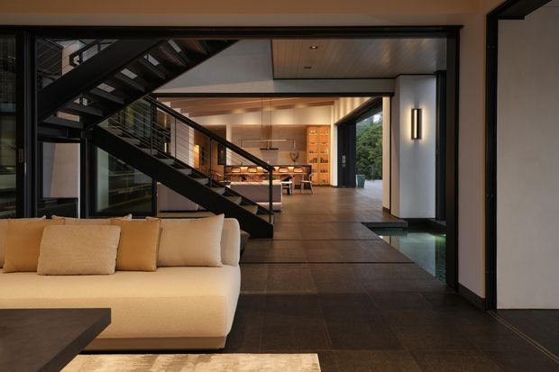 A contemporary open-plan interior with a living room in the foreground, a dramatic black metal staircase crossing the space, and a kitchen and dining area beyond. Dark large-format flooring, warm wood ceiling panels, integrated cabinetry, architectural lighting, and expansive glazing create a refined indoor-outdoor connection.