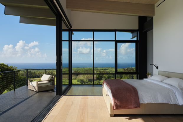 Modern bedroom opening onto a terrace, featuring floor-to-ceiling black-framed glazing, pale timber flooring and ceiling, a minimalist upholstered bed, and expansive ocean and coastal vegetation views.