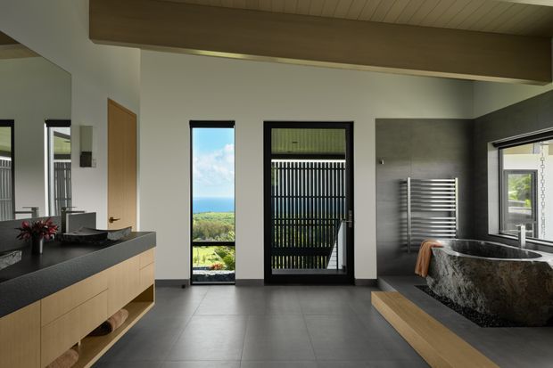 Contemporary bathroom with a freestanding dark stone bathtub, wall-mounted towel warmer, large-format grey floor tiles, and pale timber vanity cabinetry with a dark benchtop. Black-framed windows and a glazed door provide strong natural light and views toward the ocean, while a timber-lined ceiling and exposed timber beam add warmth.