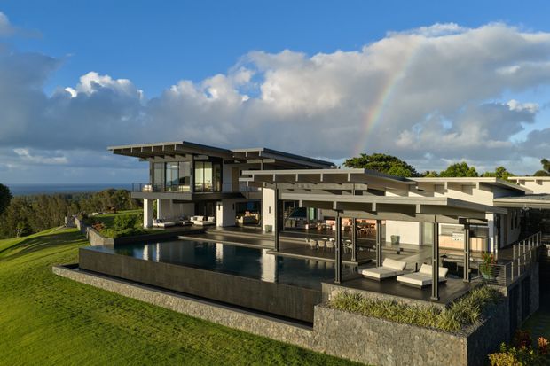 A large contemporary coastal residence viewed from outside, featuring broad flat and low-pitched roof planes, extensive floor-to-ceiling glazing, cantilevered upper volumes, covered outdoor entertaining areas, and a dark-edged infinity pool. The house is set on a landscaped sloping site with ocean visible beyond, under dramatic clouds and a rainbow.