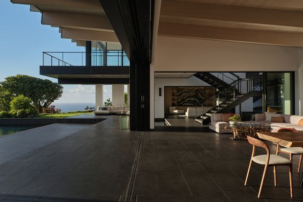A spacious contemporary open-plan indoor-outdoor living and dining area opening onto a pool terrace and landscaped coastal-looking setting. The space features dark tiled flooring, extensive glazing, black metal framing and balustrades, exposed timber soffits, a dramatic double-height void, and a staircase leading to an upper-level balcony. Neutral upholstered seating and a timber dining table furnish the living and dining zones.