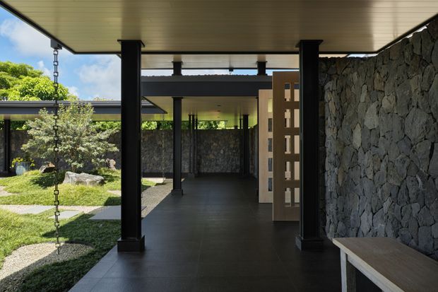 A covered architectural walkway or entry passage opening onto a landscaped courtyard. The space features a dark tiled floor, black structural posts and beams, a light-toned slatted ceiling, a rugged dark stone feature wall, a perforated timber screen door, and a simple built-in bench. Rain chains and carefully designed garden planting are visible at the courtyard edge.