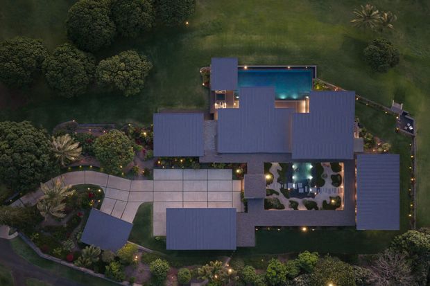 Aerial dusk view of a large, multi-wing residential property set within an extensively landscaped site. Several dark pitched roof volumes surround a central courtyard and illuminated swimming pool, with paved terraces, garden paths, mature trees, and outdoor areas visible around the building.