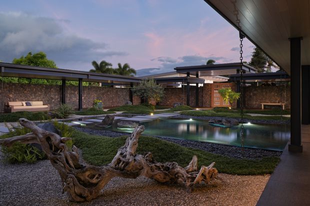 A tranquil landscaped courtyard viewed from beneath a covered outdoor structure, featuring a reflective pond with stepping stones, dark gravel, sculptural driftwood, clipped planting, stone walls, timber screening, and low modern rooflines at dusk.