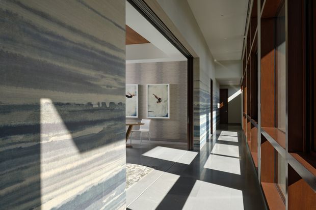 A refined contemporary interior corridor with strong linear perspective and dramatic sunlight. The hallway features a dark polished floor, pale textured stone or plaster wall surfaces, timber-framed glazed or panelled joinery along the right side, and an open doorway leading to a small dining or sitting area with framed artwork. Long rectangular shadows and clerestory-like glazing create a bright, architectural atmosphere.