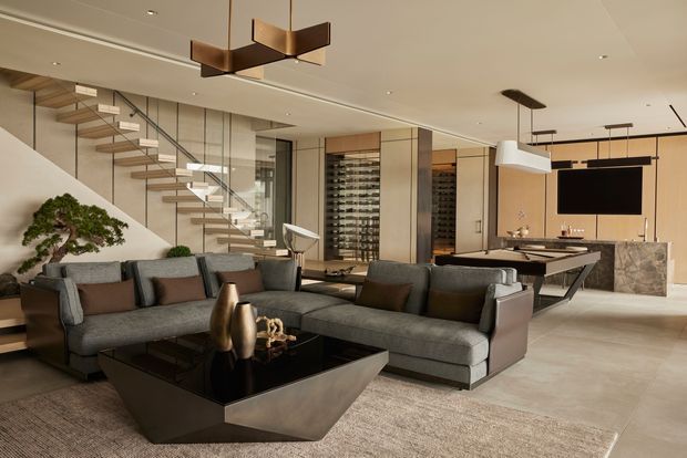 A sophisticated open-plan living room with a large grey sectional sofa, sculptural dark coffee table, neutral rug, and contemporary pendant lighting. A floating timber-and-glass staircase, extensive built-in wine storage, and a marble-clad kitchen island are visible in the background. The space has a refined, warm contemporary character with clean lines and a neutral palette.