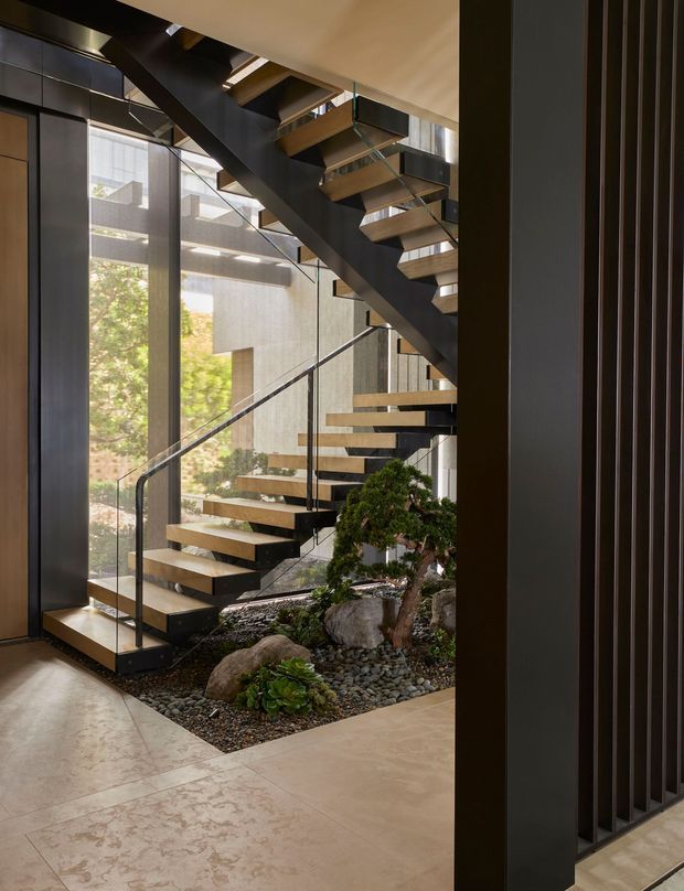 A dramatic interior stairway with floating timber treads and dark structural stringers, enclosed by tall floor-to-ceiling glazing. Clear glass balustrades, dark vertical screening, and a landscaped pebble garden with rocks and bonsai-style planting sit beneath and beside the stairs. The space has a refined contemporary character with strong natural light and a substantial open vertical volume.
