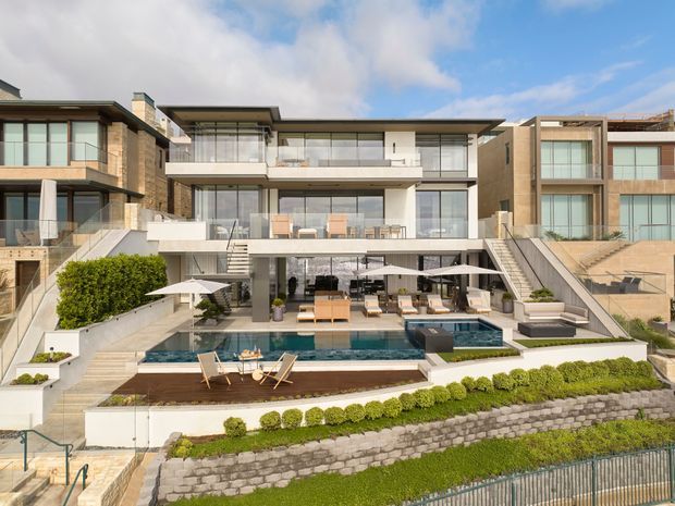 A large contemporary multi-storey residence viewed from the landscaped rear garden, with extensive floor-to-ceiling glazing, projecting balconies, pale stone or rendered walls, multiple staircases, and a stepped outdoor entertaining area. The lower level opens onto a furnished terrace beside a rectangular swimming pool, with loungers, umbrellas, and built-in seating. Terraced retaining walls and clipped shrubs frame the foreground.