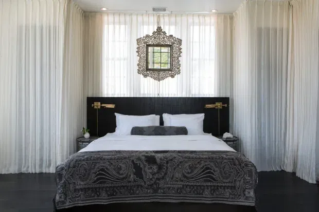 A symmetrical, refined bedroom with a centrally positioned bed, white bedding, a black upholstered or paneled headboard, and an ornate dark patterned throw. Floor-to-ceiling sheer white curtains line the walls around a large window, with an elaborate decorative mirror above the bed and brass wall-mounted reading lights on either side. The room has dark flooring and a polished, dramatic monochrome palette with metallic accents.
