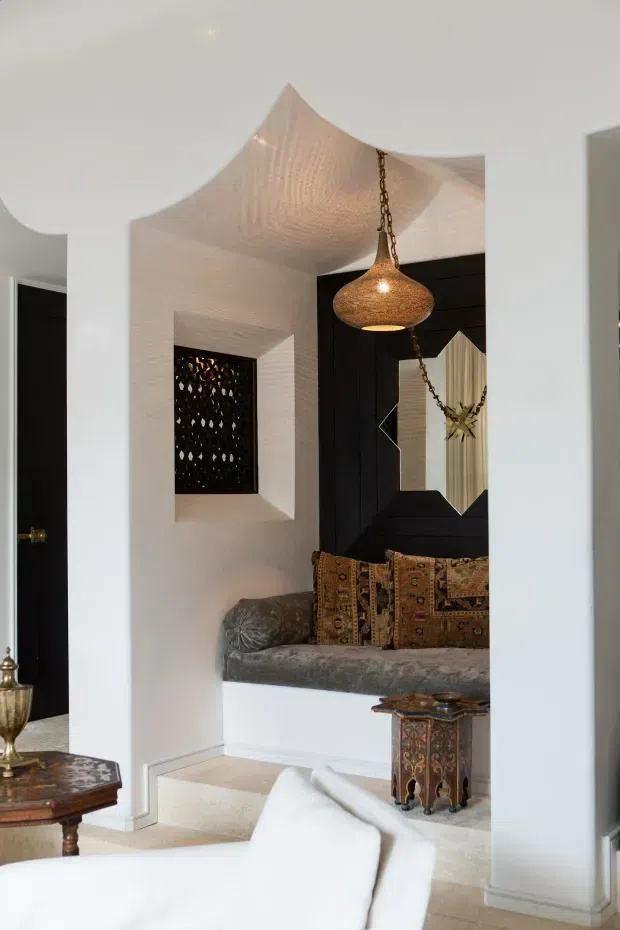 Interior view of a refined living room featuring a built-in window-seat or bench upholstered in grey velvet, patterned cushions, a large black geometric-framed mirror, and an ornate pendant light suspended from a sculptural curved white ceiling. White walls and pale flooring contrast with dark timber or painted-black joinery and richly patterned furnishings.