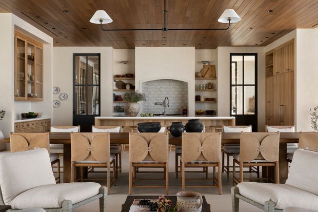 A refined open-plan kitchen and dining area viewed from the living space. A long timber dining table with upholstered dining chairs occupies the foreground, flanked by two light armchairs. The kitchen features a large island with pale cabinetry, open shelving, integrated display cabinetry, a tiled arched backsplash niche, and dark-framed glazed doors and windows. The room has a warm timber-lined ceiling with recessed downlights and suspended pendant lights, creating a balanced, inviting atmosphere.