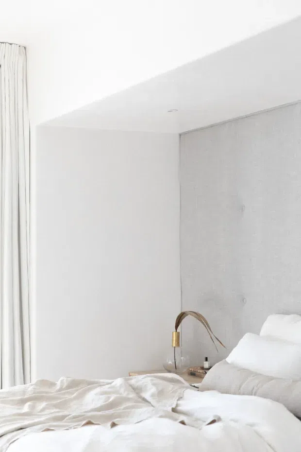 Bright, minimalist bedroom with an unmade white bed, soft white bedding, a pale textured upholstered feature wall, white painted walls and ceiling, and full-height white curtains. A small bedside table holds a simple sculptural lamp and a few items, creating a calm, airy atmosphere.