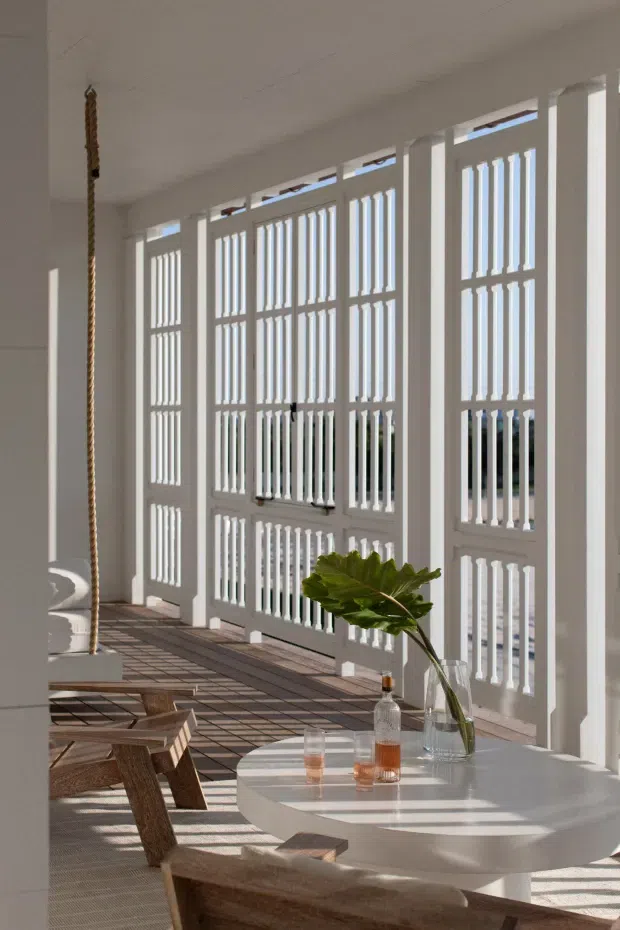 Bright, airy interior dining space with a long wall of white louvred and barred windows or shutters, casting strong linear shadows across the tiled floor. A round white dining table is paired with simple timber seating, with a tall vase and drinks on the table. The room has a relaxed coastal character and abundant daylight.