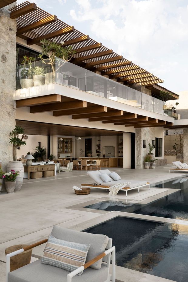 A luxurious multi-level outdoor living area centred around a dark rectangular swimming pool. The contemporary residence features a broad timber pergola with repeated slats, exposed timber beams, pale stone or plaster walls, glass balcony balustrades, and extensive indoor-outdoor glazing. A covered open-plan entertaining area includes a kitchen, dining table, bar seating, and lounge furnishings, while pale sun loungers and potted plants surround the pool.
