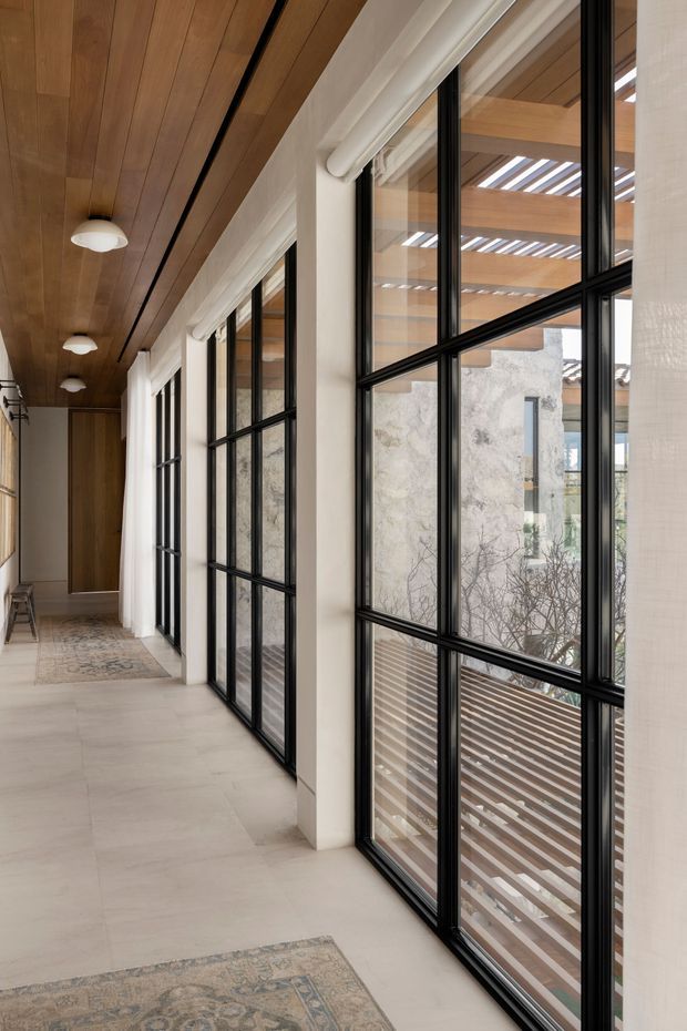 A long, light-filled interior corridor or hallway lined with tall black-framed grid windows on one side. The space has a warm timber-lined ceiling with slim black recessed detailing and small white pendant or flush-mounted lights, pale large-format flooring, white structural columns and walls, and a patterned area rug. Through the glazing, timber pergola beams and a timber deck are visible. The design has a refined contemporary character with strong indoor-outdoor connection.