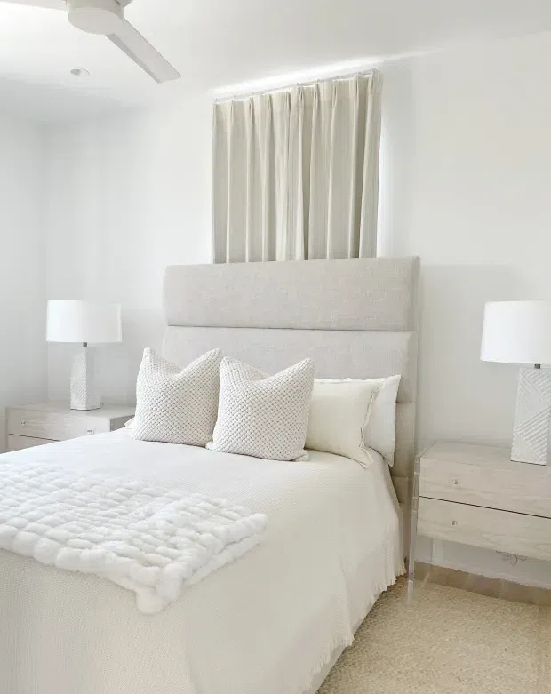 Bright, serene bedroom in a predominantly white and cream palette. The room features a large upholstered light fabric bedhead, layered white bedding and textured cushions, matching pale bedside tables with white lamps, full-height cream curtains, a white ceiling fan, and light carpeted flooring.