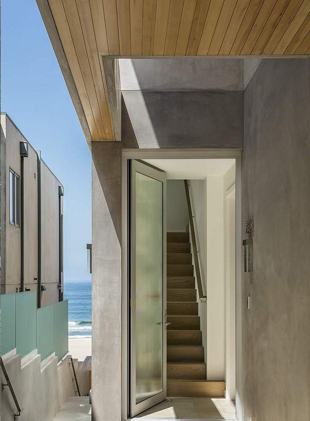 A contemporary coastal entryway viewed from outside, with a tall glazed front door opening onto a narrow stair hall. The entry features a warm timber soffit, smooth grey concrete walls, white interior walls, and a dark timber staircase with a simple handrail. A glimpse of the ocean and beach is visible beyond the adjacent passage, while neighbouring modern homes line the left side.
