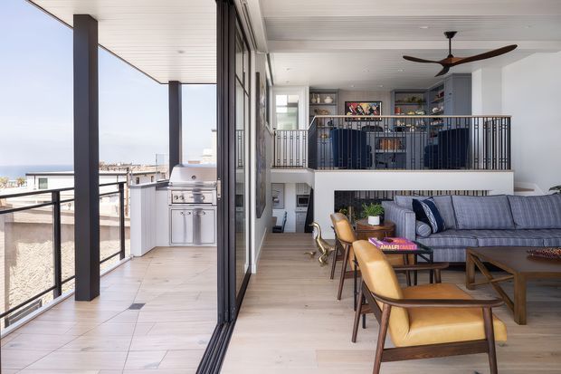 A bright contemporary open-plan living area opening onto a covered balcony or terrace. The room features a large sectional sofa, leather dining chairs, a timber dining table, a dark coffee table, and a ceiling fan. Full-height folding or sliding glazed doors connect the interior to an outdoor entertaining area with a built-in stainless-steel barbecue and outdoor cabinetry. A mezzanine-style upper level with dark metal balustrading and shelving overlooks the living space. The palette is predominantly white, light timber, grey, and black, with expansive coastal views beyond the terrace.