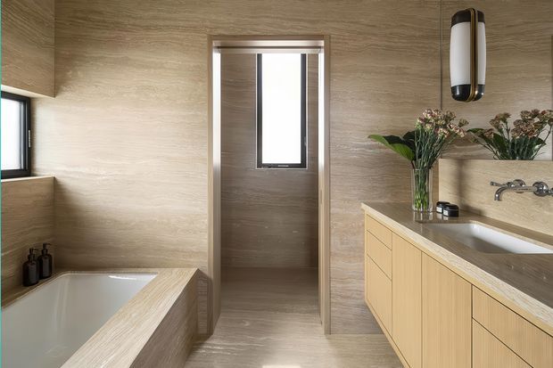 A warm, contemporary bathroom clad in light wood-grain wall and floor finishes. The room includes a partially visible built-in bathtub, a timber-look vanity with integrated basin and wall-mounted taps, narrow vertical windows, and a sculptural wall light. A doorway leads to a small enclosed area with another window. Neutral natural finishes and simple joinery create a refined, spa-like atmosphere.