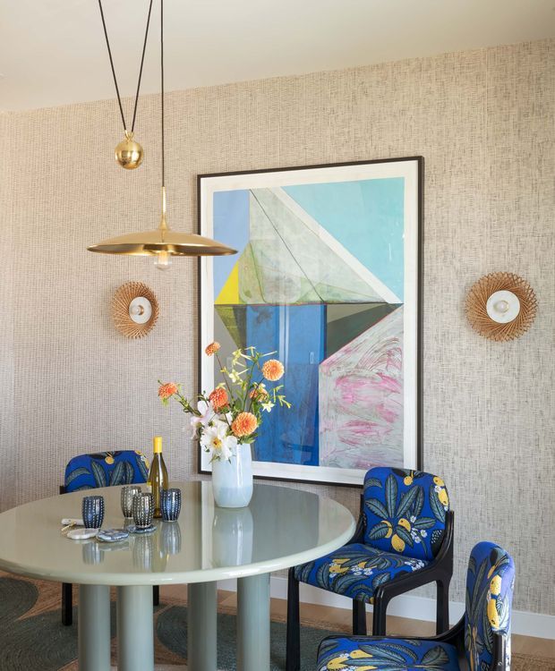 A bright, refined dining room centered on a round glass-topped dining table with sculptural pale legs and patterned blue upholstered chairs. The walls are covered in textured beige wallpaper, complemented by a large geometric abstract artwork, brass pendant lights, and matching woven sunburst wall sconces. A floral arrangement and tableware add colour and softness.
