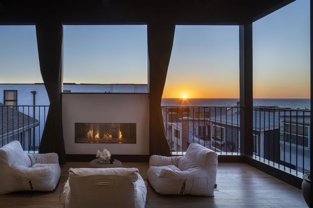 A moody, modern outdoor lounge or enclosed terrace overlooking the ocean at sunset. The space features a built-in linear fireplace set into a pale wall, dark structural framing, full-height glazing or open balcony openings, black metal balustrades, timber-look flooring, and several large white lounge chairs with a low ottoman. Coastal apartment or resort buildings are visible below and beyond the terrace.
