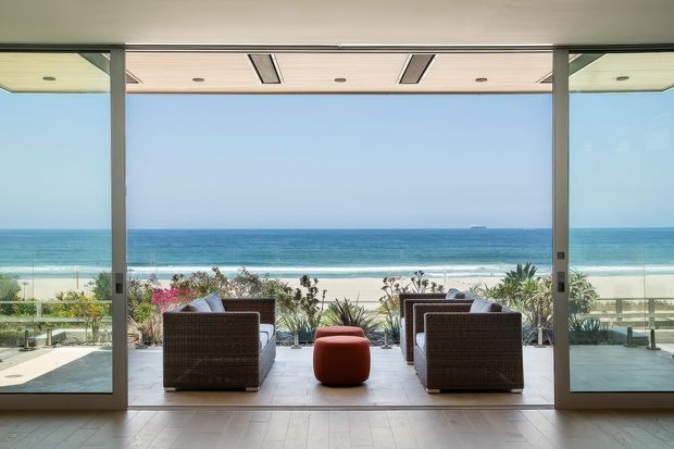 A bright, expansive indoor-outdoor living space framed by full-height sliding glass doors, opening directly onto a terrace with woven lounge chairs and a red ottoman. Beyond the landscaped edge and balustrade is a sandy beach and ocean horizon. The room has a flat ceiling with recessed linear vents and downlights, pale tiled flooring, and slim dark-framed glazing.