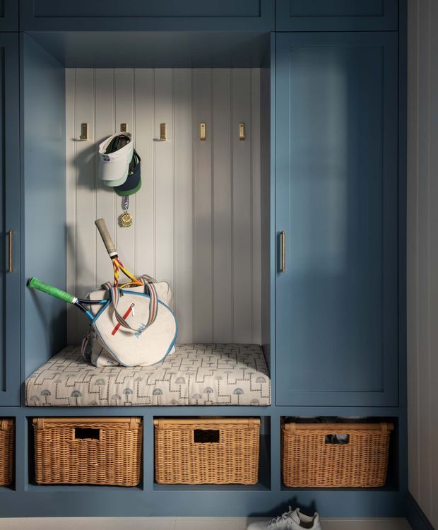 A built-in mudroom or entryway storage unit in deep blue cabinetry, with an open cubby featuring hooks, a padded patterned bench seat, sports equipment, and woven baskets in lower compartments. The space is compact, tailored, and warmly lit, with brass hardware and light vertical wall panelling.