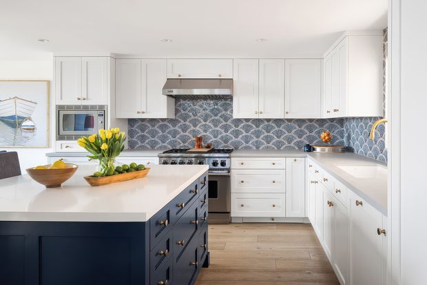 Bright, spacious kitchen with white shaker-style cabinetry, a large navy island with brass hardware, pale timber flooring, and a patterned blue-and-white scallop tile splashback. The room includes a freestanding range, stainless steel hood, integrated microwave, extensive storage, and a light stone island benchtop. Natural produce and decorative items add colour, while recessed ceiling lighting provides an even, clean illumination.