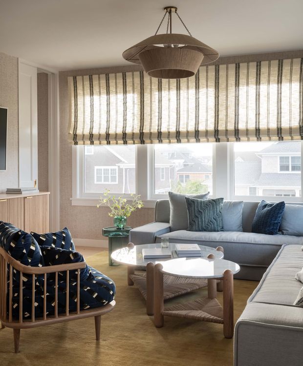 A bright, refined living room with expansive windows, woven Roman blinds, a large grey sectional sofa, patterned upholstered lounge chair, and a rounded white coffee table with woven lower shelf. A sculptural woven pendant light hangs centrally, while pale timber cabinetry, textured neutral walls, and blue patterned cushions add warmth and visual interest. The room overlooks neighbouring residential buildings.