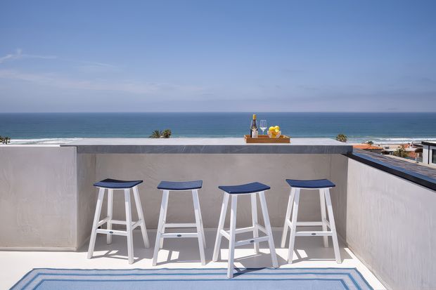 A bright rooftop terrace overlooking the ocean, with a white built-in bar counter and four white stools with dark blue seats. A small drinks setup sits on the counter, while a blue-and-white outdoor rug defines the foreground. The low parapet walls and expansive horizon create an airy coastal setting.