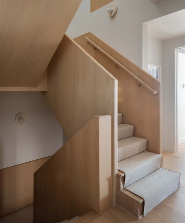 Interior view of a compact stairway with light timber panelled sides and integrated handrails. The stairs are upholstered in a pale neutral carpet, with a small landing and adjoining bright white room visible at the right. The design is clean, warm, and minimalist, with angular timber balustrade and enclosing cabinetry-like panels.
