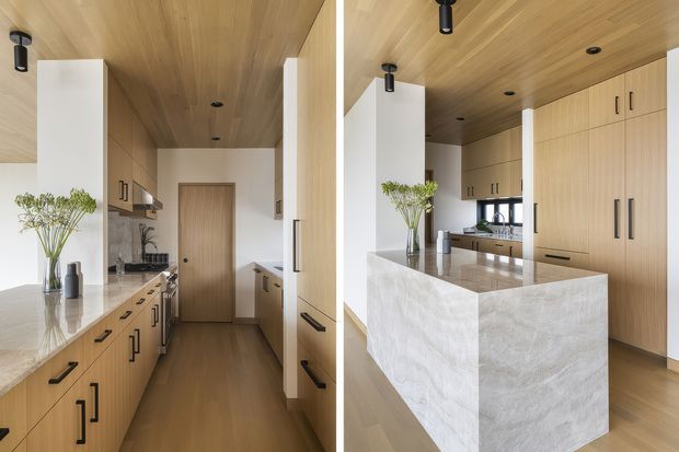A bright, contemporary galley kitchen shown from two adjoining views, with extensive light timber cabinetry, a matching timber ceiling, pale stone benchtops, and a large waterfall-edge island. Black linear hardware and recessed or track-style spot lighting provide contrast, while integrated appliances and a timber door are visible toward the rear.