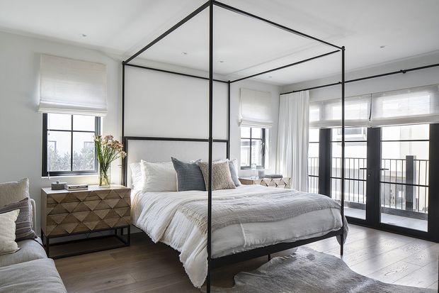 Bright, refined bedroom with a black metal four-poster canopy bed, layered white and grey bedding, and a geometric timber bedside cabinet. Large black-framed windows and glazed balcony doors bring in abundant natural light, while pale walls, light timber flooring, and sheer curtains create an airy contemporary feel.