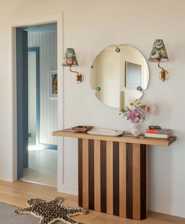 A refined entryway with a striped timber console, sculptural wall mirror, and matching decorative wall sconces. The console combines alternating warm wood tones with a slim wood top and is styled with a vase of flowers, tray, bowl, and books. Pale panelled walls, light timber flooring, a patterned animal-hide rug, and a blue-painted doorway create a warm, eclectic and elegant interior.